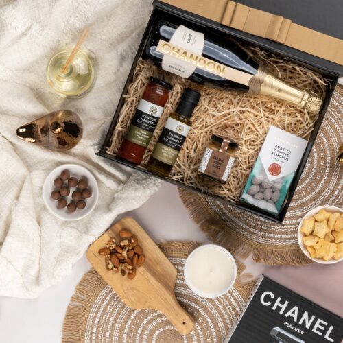An exquisite arrangement of props frames a gift hamper in this captivating flat-lay composition. The contents of the gift hamper are a thoughtfully chosen selection of delightful treats and gourmet delicacies, a true gastronomic delight. This gift hamper contains: Chutney and Pastes; Chocolate; Desert Sauce; Oil; Sparkling Wine