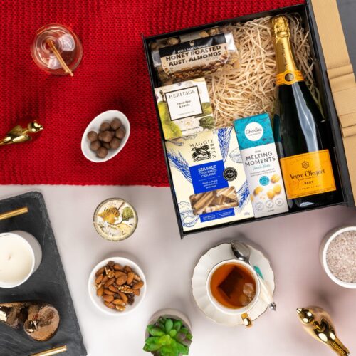 An artful arrangement of props in this captivating flat-lay image creates a sense of anticipation, as they converge towards a central gift hamper. Unveil the gift hamper to reveal a carefully selected assortment of delights, each one crafted to bring joy and enchantment. This gift hamper contains: Biscuits; Crackers; Nuts; Candles; Sparkling Wine