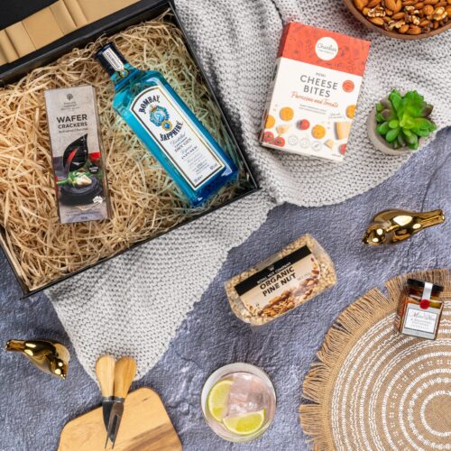 This meticulously arranged flat-lay image features a variety of props thoughtfully positioned to enhance the appeal of a gift hamper. Unbox the gift hamper to reveal a world of delightful items, each one thoughtfully selected to create a memorable occasion and leave a lasting impression. This gift hamper contains: Nuts; Crackers; Biscuits; Antipasto; Gin