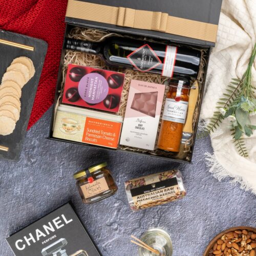 This meticulously styled flat-lay image showcases an array of props artfully positioned around a gift hamper, exuding a sense of excitement and intrigue. The gift hamper is filled with an exciting assortment of handpicked products, ready to be savored and enjoyed. This gift hamper contains: Red Wine; Mustards and Dressings; Nuts; Biscuits; Chocolate; Antipasto