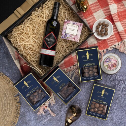 This striking flat-lay image features a curated selection of props that converge around a gift hamper, speaking of generosity and thoughtfulness. The gift hamper holds an enchanting array of carefully selected surprises, each one contributing to a memorable and magical experience, filling the recipient's heart with wonder. This gift hamper contains: Chocolate; Red Wine; Candles
