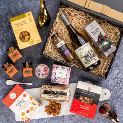 This captivating flat-lay composition entices the viewer with a carefully arranged assortment of props. The gift hamper is filled with an exciting assortment of handpicked products, carefully chosen to evoke delight and satisfaction, creating a moment of pure indulgence and pure pleasure. This gift hamper contains: White Wine; Mustards and Dressings; Chutney and Pastes; Candles; Nuts; Biscuits; Crackers
