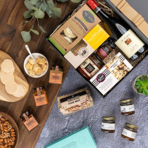 This captivating flat-lay image features a carefully curated collection of props arranged artfully around a gift hamper. The gift hamper holds an enticing mix of carefully selected products and gourmet delights. This gift hamper contains: Mustards and Dressings; Chutney and Pastes; Biscuits; Red Wine; Crackers; Jam; Nuts