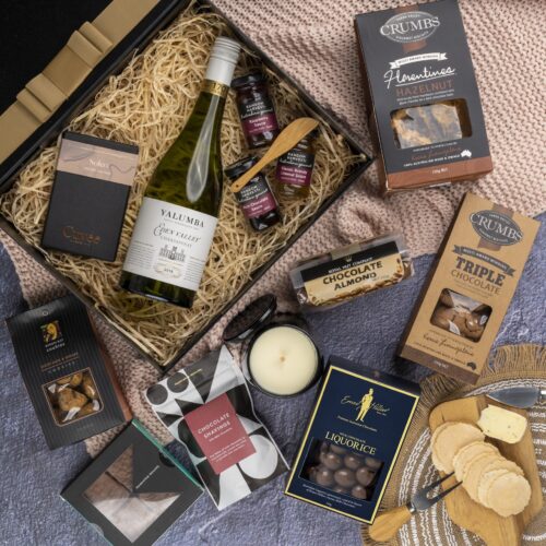 A meticulously arranged collage of props forms a picturesque backdrop for this flat-lay image, enhancing the allure of a gift hamper. The gift hamper holds an enchanting array of carefully selected surprises, guaranteed to leave a lasting impression. This gift hamper contains: Desert Sauce; Biscuits; Chocolate; Drinking Chocolate; White Wine; Candles