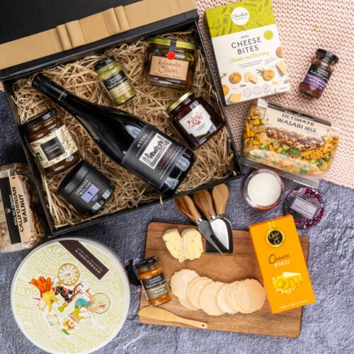 A symphony of colors and textures unfolds in this captivating flat-lay photograph, as an array of props encircle a gift hamper. Inside the gift hamper, you'll discover a carefully curated collection of exquisite items, each one a testament to luxury and elegance, ensuring a remarkable gift-giving experience that will be cherished and treasured, creating moments of pure happiness and pure indulgence. This gift hamper contains: Candles; Cheese Board; Chocolate; Jam; Mustards and Dressings; Biscuits; Nuts; Antipasto; Red Wine