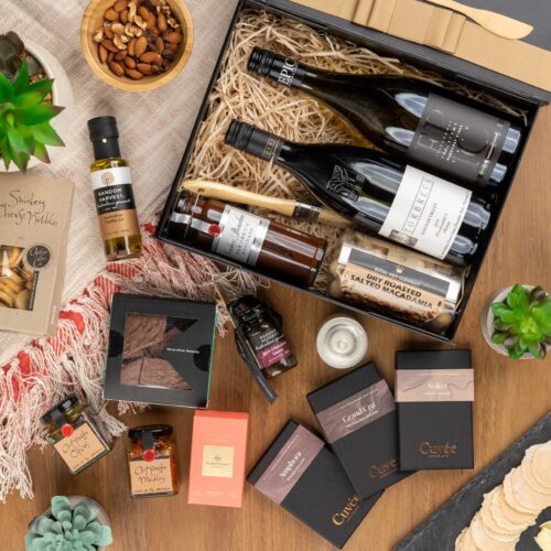 This visually dynamic flat-lay composition showcases a blend of props skillfully positioned around a gift hamper. The contents of the gift hamper promise a journey of indulgence and discovery, with each item more delightful than the last. This gift hamper contains: Antipasto; White Wine; Crackers; Chocolate; Nuts; Red Wine; Candles; Oil; Mustards and Dressings
