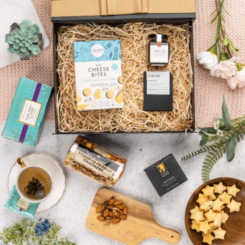This thoughtfully crafted flat-lay image features an interplay of textures and shapes, as props converge around a gift hamper. The contents of the gift hamper promise a journey of indulgence and discovery, with each item carefully selected to bring joy and happiness, making every moment special and unforgettable. This gift hamper contains: Biscuits; Jam; Nuts; Chocolate; Tea (Black)