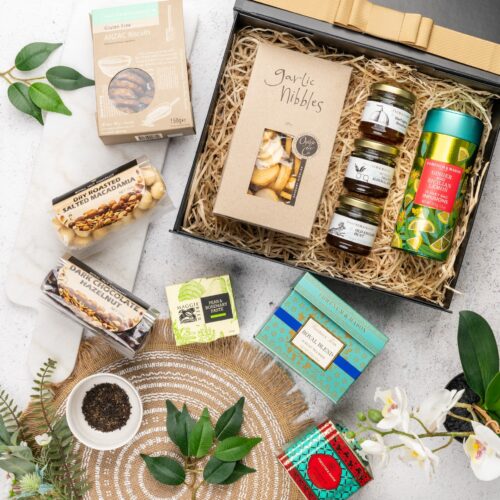 In this carefully composed flat-lay photograph, props of varying sizes and shapes are placed with precision, forming a beautiful frame for a gift hamper. Discover a captivating collection of items within the gift hamper, each one thoughtfully chosen to create a truly remarkable experience, taking the recipient on a journey of delight. This gift hamper contains: Biscuits; Chutney and Pastes; Crackers; Tea; Jam; Nuts; Chocolate