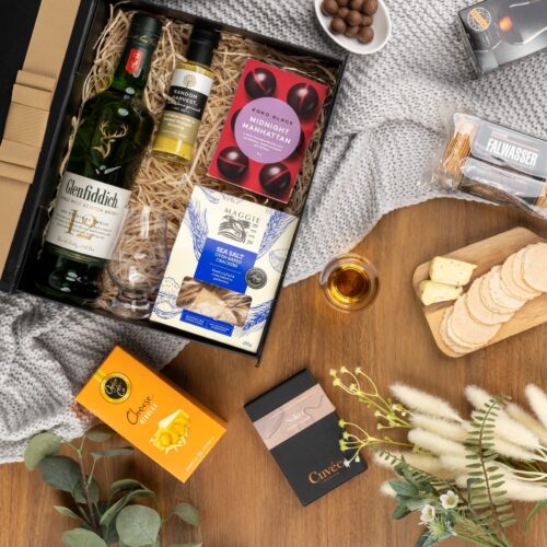 This visually captivating flat-lay composition showcases an interplay of light and shadow as an assortment of props encircle a gift hamper. Inside the gift hamper, you'll discover a carefully curated collection of exquisite items, each one a testament to luxury and elegance. This gift hamper contains: Chocolate; Whisky; Mustards and Dressings; Biscuits; Crackers