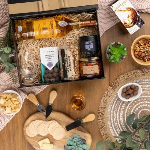 A visual feast awaits in this flat-lay photograph, where a melange of props frames a gift hamper, hinting at the exciting possibilities. The contents of the gift hamper are a collection of delightful treats and luxurious indulgences. This gift hamper contains: Whisky; Antipasto; Chocolate; Nuts