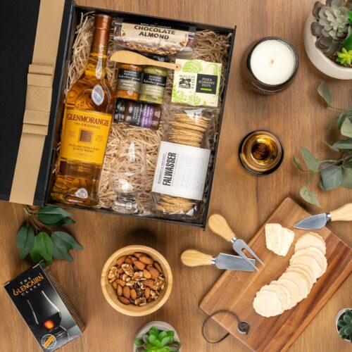 A meticulously arranged collage of props forms a picturesque backdrop for this flat-lay image, enhancing the allure of a gift hamper. The contents of the gift hamper are a thoughtfully chosen selection of delightful treats and gourmet delicacies, a true gastronomic delight that will tantalize the taste buds and please the senses, a gift that satisfies both the body and the soul. This gift hamper contains: Biscuits; Whisky; Candles; Chocolate; Chutney and Pastes; Mustards and Dressings