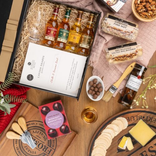 In this visually striking flat-lay image, an assortment of props is skillfully positioned to enhance the allure of a gift hamper. Open the gift hamper to uncover a meticulously chosen selection of items, each one contributing to a truly delightful experience. This gift hamper contains: Nuts; Mustards and Dressings; Chocolate; Whisky; Biscuits