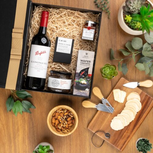 This captivating flat-lay image features a carefully curated collection of props arranged artfully around a gift hamper. The contents of the gift hamper are a thoughtfully chosen selection of delightful treats and gourmet delicacies, a true gastronomic delight that will tantalize the taste buds and please the senses. This gift hamper contains: Chocolate; Jam; Red Wine; Antipasto; Crackers