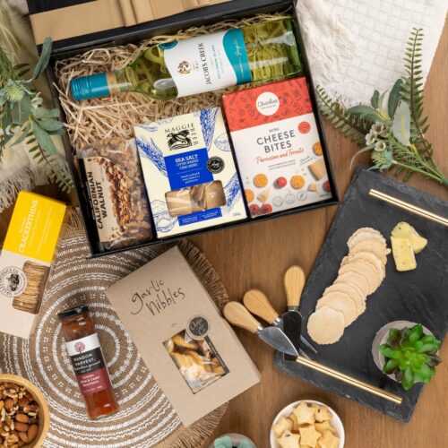 In this carefully composed flat-lay photograph, props of varying sizes and shapes are placed with precision, forming a beautiful frame for a gift hamper. This thoughtfully crafted gift hamper contains a variety of wonderful surprises. This gift hamper contains: Mustards and Dressings; White Wine; Crackers; Biscuits; Nuts