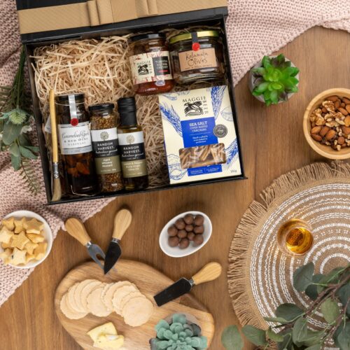 In this captivating flat-lay image, an array of carefully selected props encircles a gift hamper, creating a visual narrative. Discover a carefully curated assortment of items nestled within the gift hamper, each one more delightful than the last. This gift hamper contains: Crackers; Chutney and Pastes; Antipasto; Mustards and Dressings; Oil