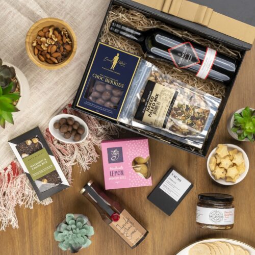 This visually arresting flat-lay image showcases a meticulously styled collection of props, expertly arranged around a gift hamper. The gift hamper is filled with an exciting variety of handpicked items, perfect for every occasion. This gift hamper contains: Red Wine; Jam; Biscuits; Desert Sauce; Chocolate; Nuts