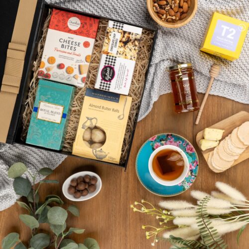 In this visually striking flat-lay composition, an assortment of props is elegantly arranged around a gift hamper, evoking a sense of luxury and sophistication. Discover a captivating collection of items within the gift hamper, each one thoughtfully chosen to create a truly remarkable experience, taking the recipient on a journey of delight and surprise, a gift that brings a sense of wonder and awe. This gift hamper contains: Biscuits; Honey; Teaware; Tea; Tea (Black)
