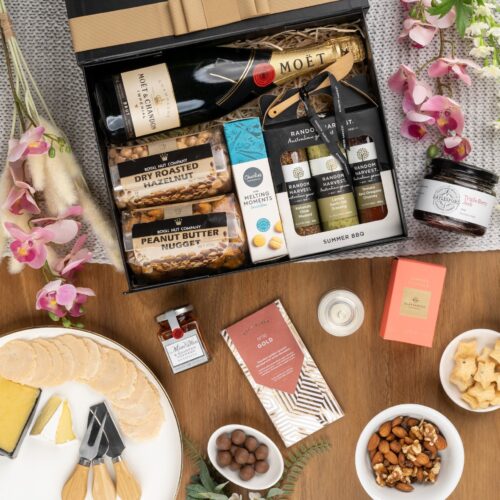 In this visually stunning flat-lay photograph, an assembly of props creates a visual narrative, highlighting the beauty and elegance of a gift hamper. The gift hamper holds an enchanting array of carefully chosen surprises, designed to bring joy and delight. This gift hamper contains: Chocolate; Jam; Biscuits; Antipasto; Sparkling Wine; Nuts; Chutney and Pastes; Candles