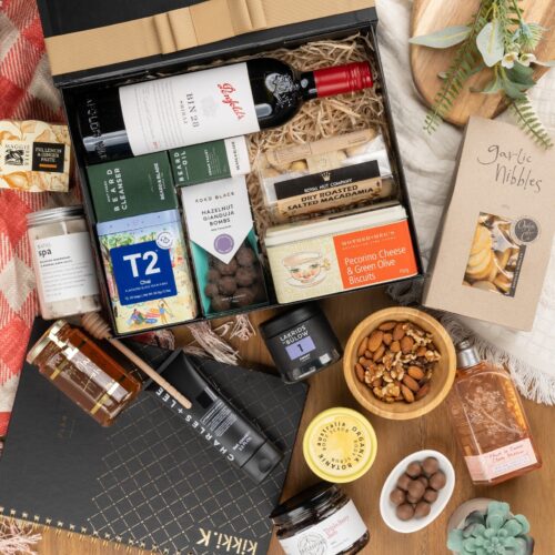 This captivating flat-lay image features a carefully curated collection of props arranged artfully around a gift hamper. Inside the gift hamper, you'll find an exquisite selection of carefully chosen items, each one a delightful surprise. This gift hamper contains: Tea (Chai); Jam; Chutney and Pastes; Red Wine; Chocolate; Skincare; Honey; Crackers; Nuts; Planners; Biscuits