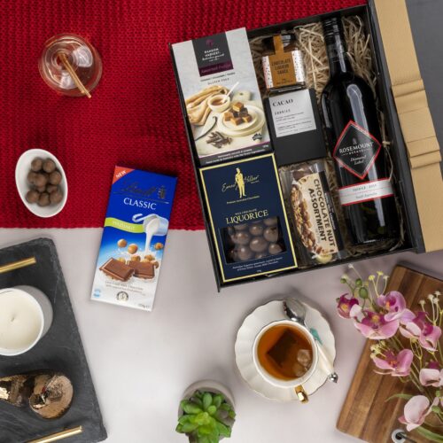 This visually dynamic flat-lay composition showcases a blend of props skillfully positioned around a gift hamper. Open the gift hamper to uncover a meticulously chosen selection of items, each one contributing to a truly delightful experience, leaving the recipient in awe and filled with gratitude. This gift hamper contains: Chocolate; Desert Sauce; Red Wine