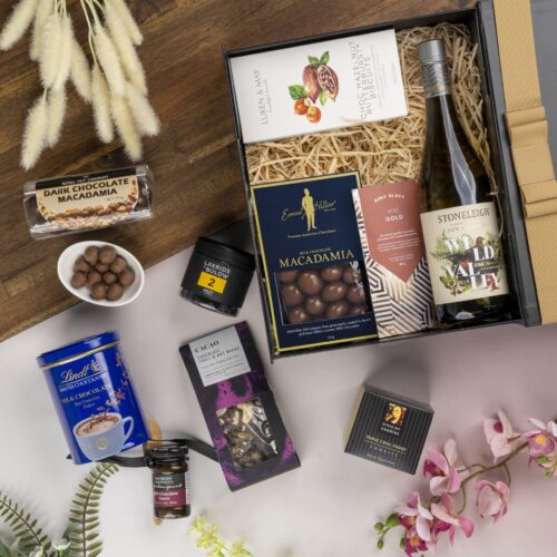 This thoughtfully crafted flat-lay image features an interplay of textures and shapes, as props converge around a gift hamper. Inside the gift hamper, you'll find an exquisite selection of carefully chosen items, each one a delightful surprise. This gift hamper contains: White Wine; Chocolate; Biscuits; Drinking Chocolate