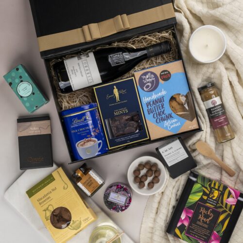 This striking flat-lay image features a curated selection of props that converge around a gift hamper, speaking of generosity and thoughtfulness. Open the gift hamper to uncover a meticulously chosen selection of items, each one contributing to a truly delightful experience, leaving the recipient in awe and filled with gratitude. This gift hamper contains: Drinking Chocolate; Biscuits; Red Wine; Chocolate; Desert Sauce; Candles