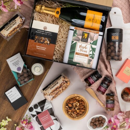 In this visually stunning flat-lay photograph, an assembly of props creates a visual narrative, highlighting the beauty and elegance of a gift hamper. Open the gift hamper to reveal a delightful surprise waiting to be savored, each item carefully selected to create a moment of pure bliss and bring a smile to the recipient's face, capturing their heart. This gift hamper contains: Desert Sauce; Chocolate; Sparkling Wine; Candles; Biscuits; Drinking Chocolate; Nuts
