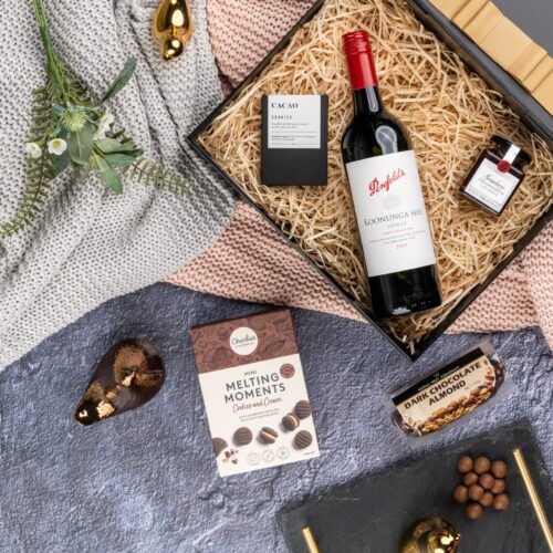 This striking flat-lay image features a curated selection of props that converge around a gift hamper, speaking of generosity and thoughtfulness. The gift hamper holds an enchanting array of carefully selected surprises, each one contributing to a memorable and magical experience. This gift hamper contains: Jam; Red Wine; Chocolate; Biscuits