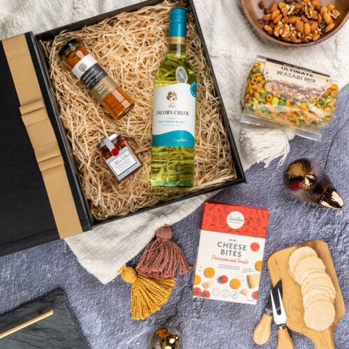 In this flat-lay photograph, a beautifully arranged assortment of props surrounds a gift hamper, creating an eye-catching composition. Inside the gift hamper, you'll find a curated collection of wonderful items, each one chosen to bring joy and happiness. This gift hamper contains: White Wine; Antipasto; Biscuits; Mustards and Dressings; Nuts