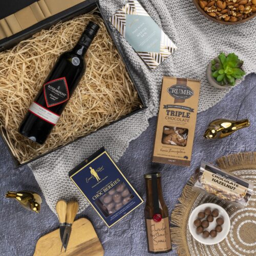 This captivating flat-lay image features a carefully curated collection of props arranged artfully around a gift hamper. The gift hamper holds an enchanting array of carefully selected surprises, guaranteed to leave a lasting impression. This gift hamper contains: Chocolate; Red Wine; Biscuits; Desert Sauce