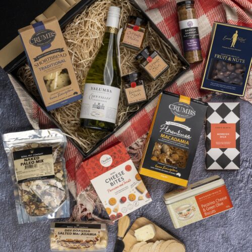 This visually arresting flat-lay image showcases a meticulously styled collection of props, expertly arranged around a gift hamper. Unbox the gift hamper to reveal a world of delightful items, each one thoughtfully selected to create a memorable occasion and leave a lasting impression, a true celebration of happiness and love, capturing the essence of joy. This gift hamper contains: Chutney and Pastes; Nuts; Biscuits; Chocolate; Antipasto; White Wine