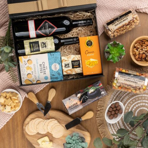 This meticulously styled flat-lay image showcases an array of props artfully positioned around a gift hamper, exuding a sense of excitement and intrigue. Open the gift hamper to uncover a meticulously chosen selection of items, each one contributing to a truly delightful experience, leaving the recipient in awe and filled with gratitude, a gift that exceeds all expectations and touches the soul. This gift hamper contains: Oil; Crackers; Biscuits; Nuts; Chutney and Pastes; Red Wine