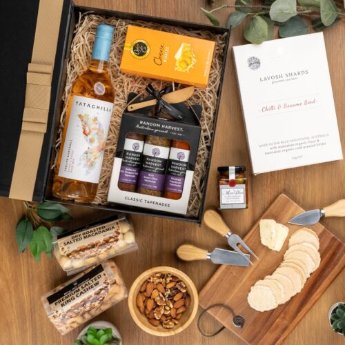 This meticulously styled flat-lay image showcases an array of props artfully positioned around a gift hamper, exuding a sense of excitement and intrigue. Open the gift hamper to uncover a meticulously chosen selection of items, carefully arranged to bring delight. This gift hamper contains: Chutney and Pastes; Antipasto; Rose Wine; Nuts; Biscuits
