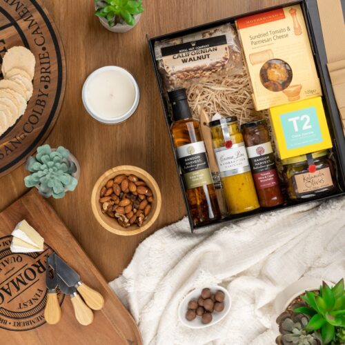 A symphony of colors and textures unfolds in this captivating flat-lay photograph, as an array of props encircle a gift hamper. The contents of the gift hamper promise a delightful and unforgettable experience. This gift hamper contains: Antipasto; Crackers; Mustards and Dressings; Tea (Green); Oil; Nuts