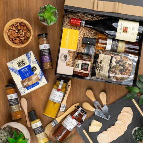 In this visually striking flat-lay composition, an assortment of props is elegantly arranged around a gift hamper, evoking a sense of luxury and sophistication. Discover a captivating collection of items within the gift hamper, carefully chosen to create a truly unforgettable experience. This gift hamper contains: Mustards and Dressings; Nuts; Crackers; Red Wine; Chutney and Pastes; Oil