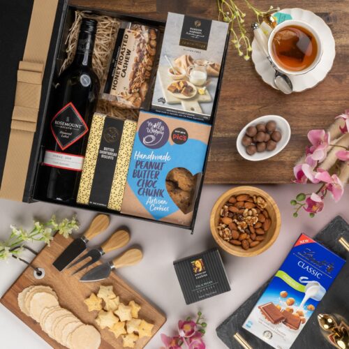 In this visually striking flat-lay image, an assortment of props is skillfully positioned to enhance the allure of a gift hamper. Unveil the gift hamper to reveal a carefully selected assortment of delights, each one crafted to bring joy and enchantment, allowing the recipient to immerse in a world of happiness and wonder. This gift hamper contains: Biscuits; Nuts; Chocolate; Red Wine