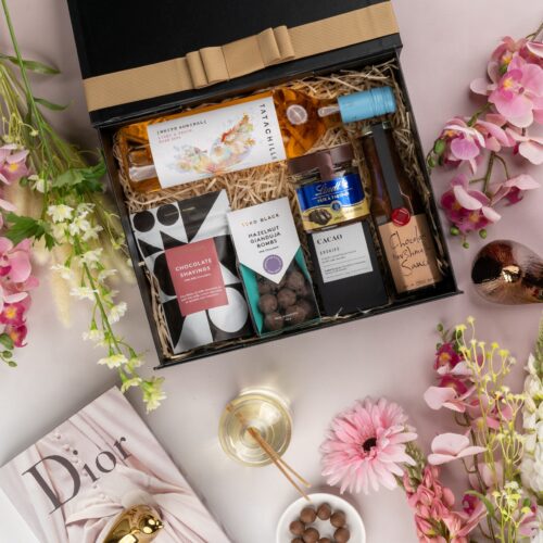 In this visually striking flat-lay image, an assortment of props is skillfully positioned to enhance the allure of a gift hamper. Inside the gift hamper, you'll discover a carefully curated collection of exquisite items, each one a testament to luxury and elegance, ensuring a remarkable gift-giving experience. This gift hamper contains: Drinking Chocolate; Chocolate; Desert Sauce; Rose Wine