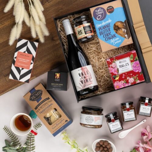 This visually captivating flat-lay composition showcases an interplay of light and shadow as an assortment of props encircle a gift hamper. The contents of the gift hamper promise a delightful and unforgettable experience. This gift hamper contains: Teaware; Biscuits; Jam; Red Wine; Chocolate