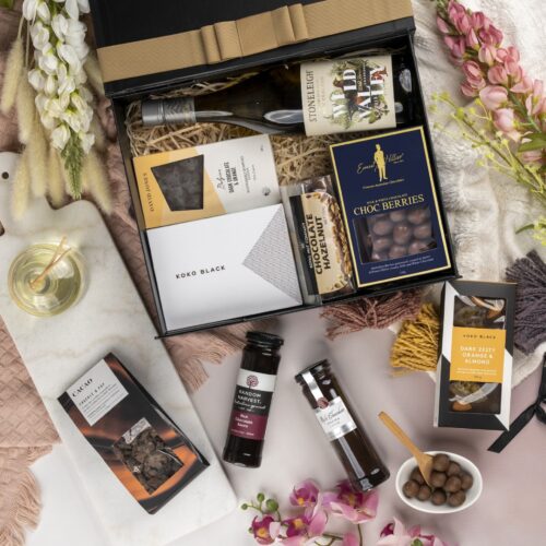 This thoughtfully crafted flat-lay image features an interplay of textures and shapes, as props converge around a gift hamper. Inside the gift hamper, you'll discover a carefully curated collection of exquisite items, each one a true delight. This gift hamper contains: White Wine; Chocolate; Desert Sauce