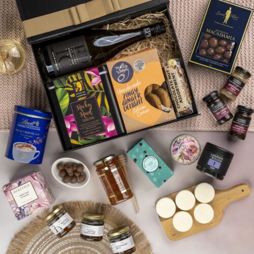 This visually dynamic flat-lay composition showcases a blend of props skillfully positioned around a gift hamper. Open the gift hamper to reveal a delightful surprise waiting to be savored, each item carefully selected to create a moment of pure bliss. This gift hamper contains: Biscuits; Desert Sauce; Honey; White Wine; Drinking Chocolate; Jam; Candles; Chocolate