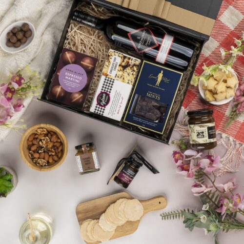 This visually dynamic flat-lay composition showcases a blend of props skillfully positioned around a gift hamper. Explore the gift hamper to find a delightful assortment of surprises, guaranteed to bring joy and excitement. This gift hamper contains: Chocolate; Red Wine; Desert Sauce; Biscuits