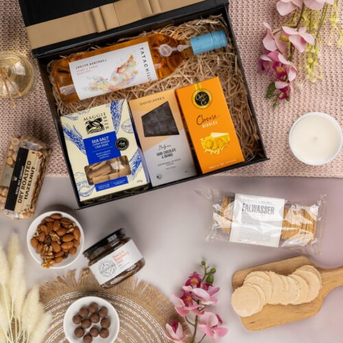 A carefully curated selection of props forms a picturesque frame around a gift hamper in this mesmerizing flat-lay photograph. The contents of the gift hamper are a carefully chosen selection of delightful treats and luxurious surprises. This gift hamper contains: Jam; Rose Wine; Biscuits; Crackers; Chocolate; Nuts
