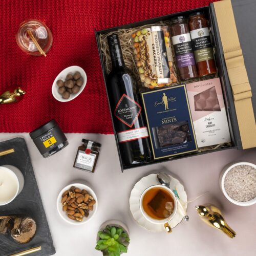 A meticulously arranged collage of props forms a picturesque backdrop for this flat-lay image, enhancing the allure of a gift hamper. Unbox the gift hamper to reveal a world of delightful items, each one thoughtfully selected to create a memorable occasion and leave a lasting impression. This gift hamper contains: Nuts; Chutney and Pastes; Chocolate; Red Wine; Jam