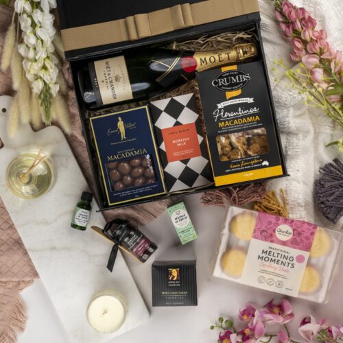 In this carefully composed flat-lay photograph, props of varying sizes and shapes are placed with precision, forming a beautiful frame for a gift hamper. Inside the gift hamper, you'll be delighted to find an assortment of delightful goodies and decadent treats. This gift hamper contains: Chocolate; Sparkling Wine; Biscuits; Essential Oil; Candles