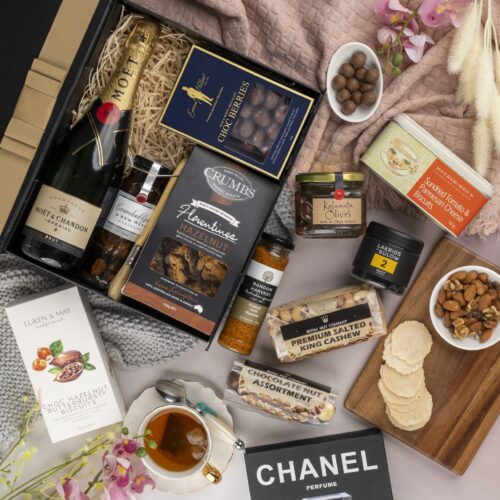 In this captivating flat-lay image, an array of carefully selected props encircles a gift hamper, creating a visual narrative. Discover a captivating collection of items within the gift hamper, each one thoughtfully chosen to create a truly remarkable experience, taking the recipient on a journey of delight. This gift hamper contains: Chocolate; Biscuits; Mustards and Dressings; Antipasto; Nuts; Sparkling Wine