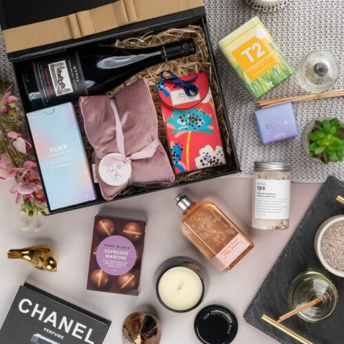 This visually captivating flat-lay composition showcases an interplay of light and shadow as an assortment of props encircle a gift hamper. Open the gift hamper to reveal a delightful surprise waiting to be enjoyed, a perfect gift for any recipient. This gift hamper contains: Red Wine; Candles; Bath And Shower Gel; Soap; Tea (Green); Pillow; Chocolate; Diffuser ; Skincare