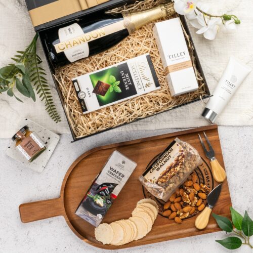 In this flat-lay photograph, a beautifully arranged assortment of props surrounds a gift hamper, creating an eye-catching composition. Open the gift hamper to uncover a meticulously chosen selection of items, each one contributing to a truly delightful experience, leaving the recipient in awe and filled with gratitude. This gift hamper contains: Nuts; Hand And Nail Cream; Desert Sauce; Sparkling Wine; Crackers; Chocolate