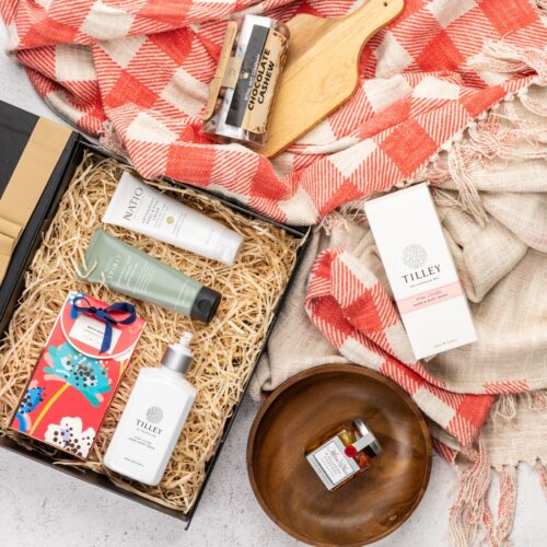 This thoughtfully crafted flat-lay image features an interplay of textures and shapes, as props converge around a gift hamper. Open the gift hamper to reveal a delightful surprise waiting to be savored, each item carefully selected to create a moment of pure bliss and bring a smile to the recipient's face, capturing their heart. This gift hamper contains: Antipasto; Chocolate; Bath And Shower Gel; Hand Cream; Hand And Body Wash; Skincare