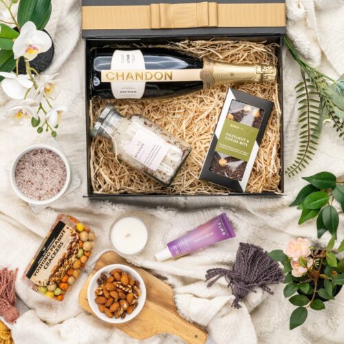 In this visually stunning flat-lay photograph, an assembly of props creates a visual narrative, highlighting the beauty and elegance of a gift hamper. Unbox the gift hamper to reveal a world of delightful items, each one thoughtfully selected to create a memorable occasion and leave a lasting impression. This gift hamper contains: Bath And Shower Gel; Candles; Nuts; Hand Cream; Sparkling Wine; Chocolate