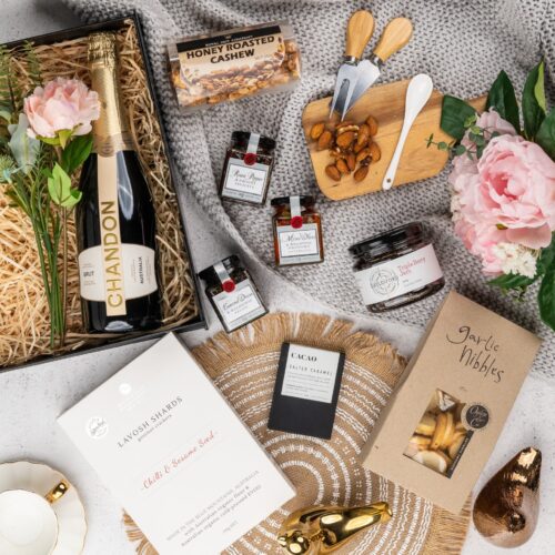 In this captivating flat-lay image, an array of carefully selected props encircles a gift hamper, creating a visual narrative. Discover a captivating collection of items within the gift hamper, each one chosen with care and thoughtfulness. This gift hamper contains: Biscuits; Nuts; Chocolate; Jam; Sparkling Wine; Mustards and Dressings